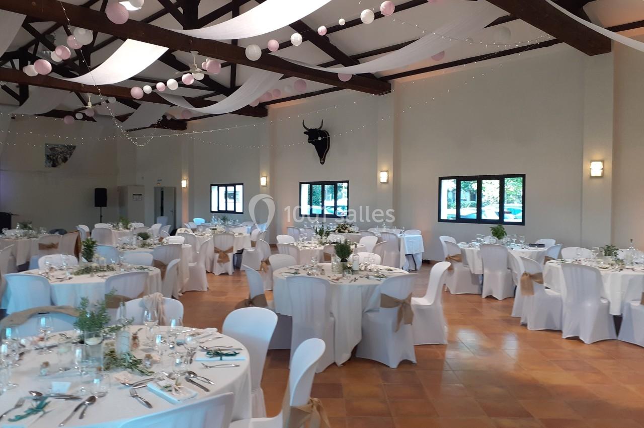 Salle de réception décorée pour un événement, avec tables rondes dressées, nappes blanches et guirlandes suspendues.