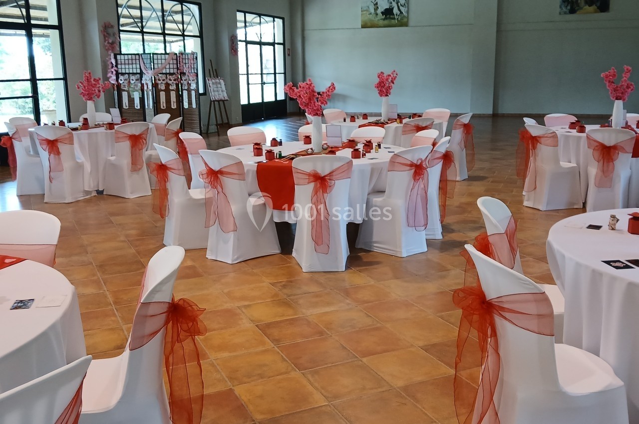 Salle de réception décorée avec des nappes blanches et des rubans rouges, tables ornées de centres de table fleuris.