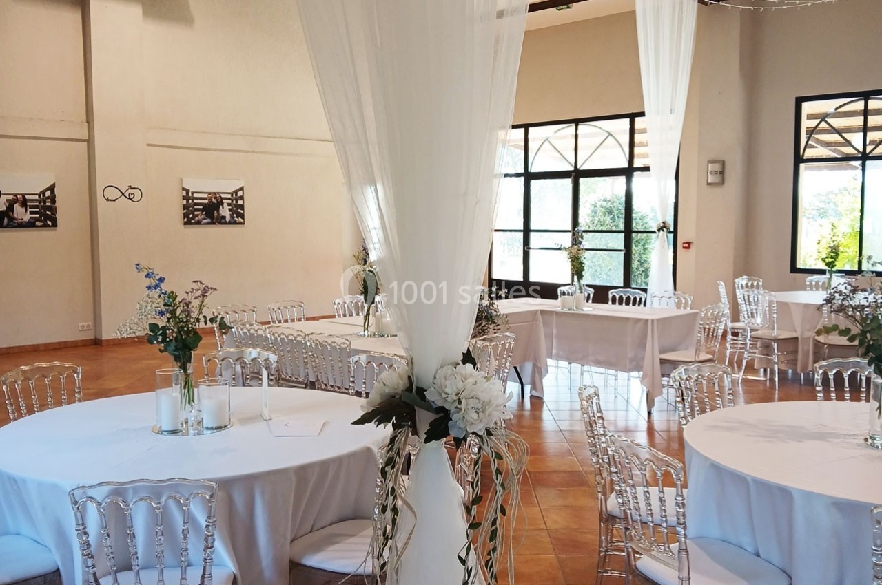 Salle de réception lumineuse avec tables rondes dressées, chaises transparentes et décoration florale élégante.
