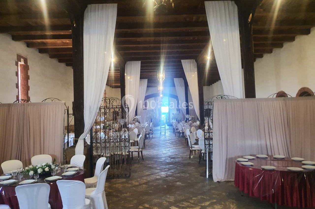 Salle de réception avec tables dressées, chaises blanches et rideaux suspendus, éclairée par des lustres.
