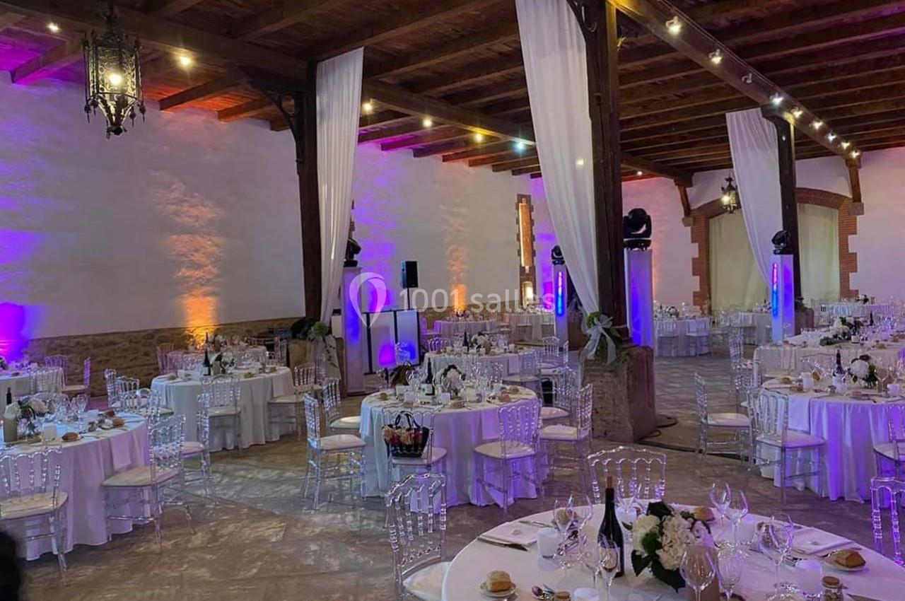 Salle de réception élégante avec tables rondes dressées, chaises transparentes et éclairage tamisé.