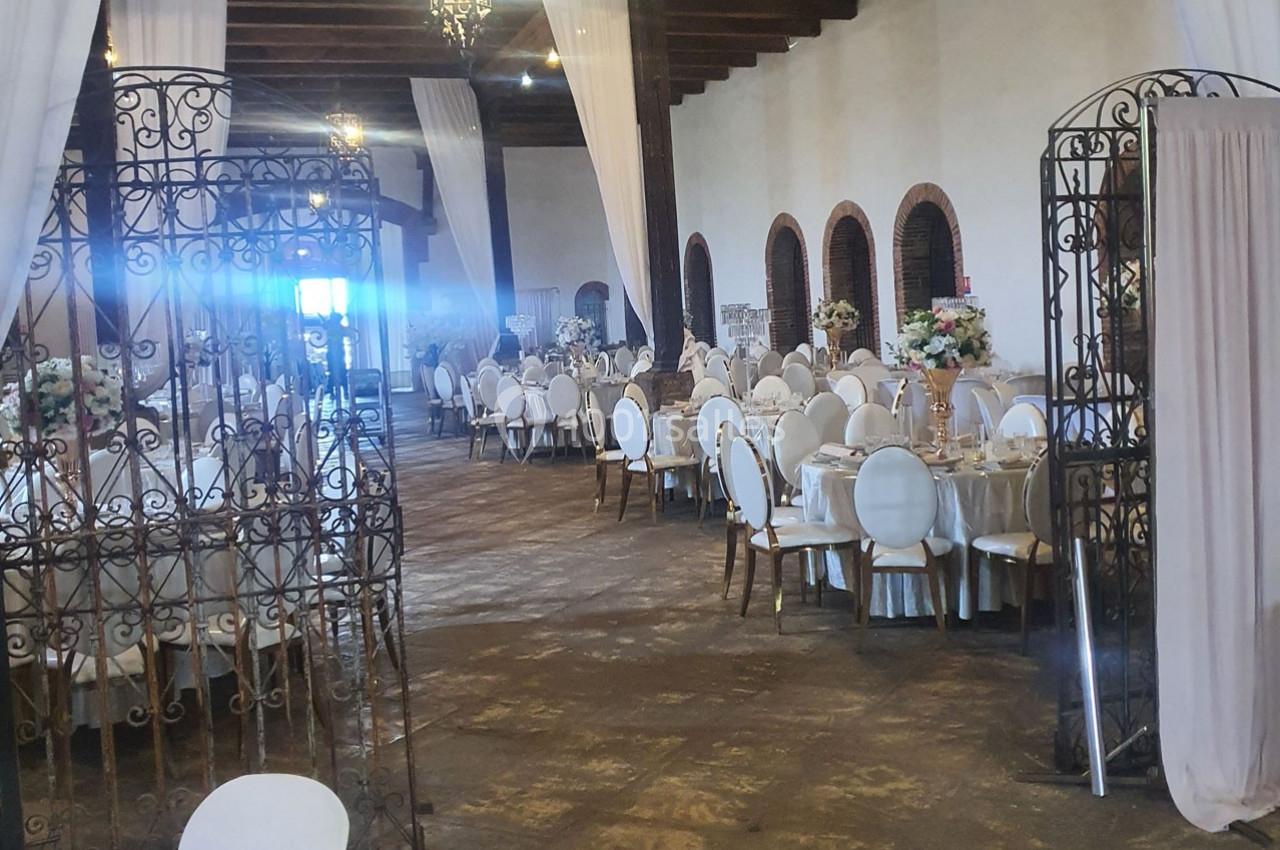 Salle de réception élégante avec tables rondes dressées, chaises blanches et décorations florales, éclairée par des lustres.