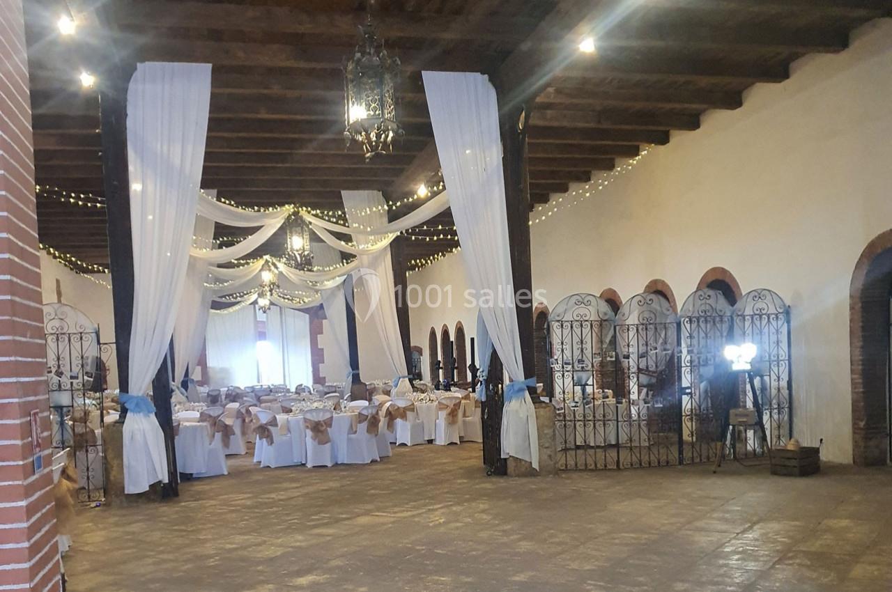 Salle de réception décorée avec des tables dressées, des voilages blancs suspendus et des guirlandes lumineuses.