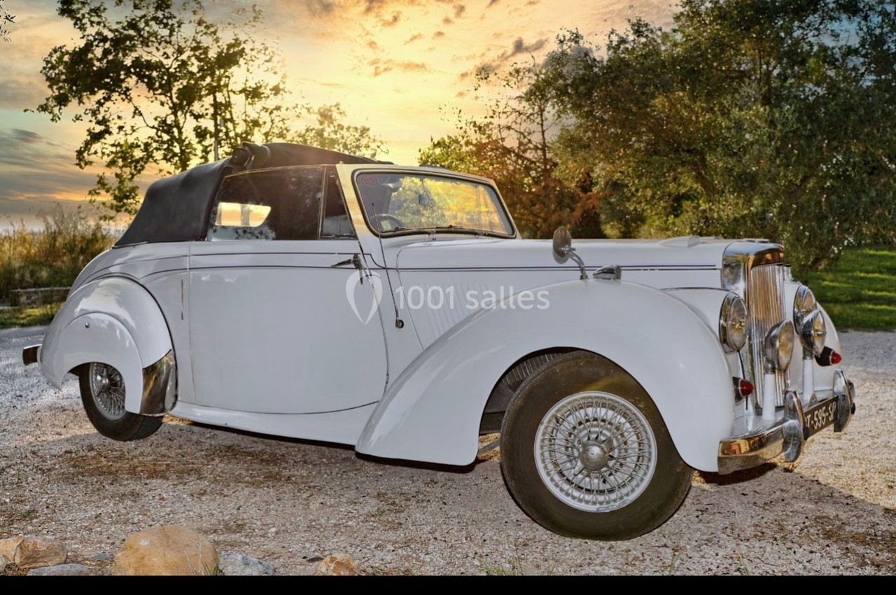 Voiture ancienne blanche décapotable stationnée sur un chemin gravillonné, avec un coucher de soleil en arrière-plan.