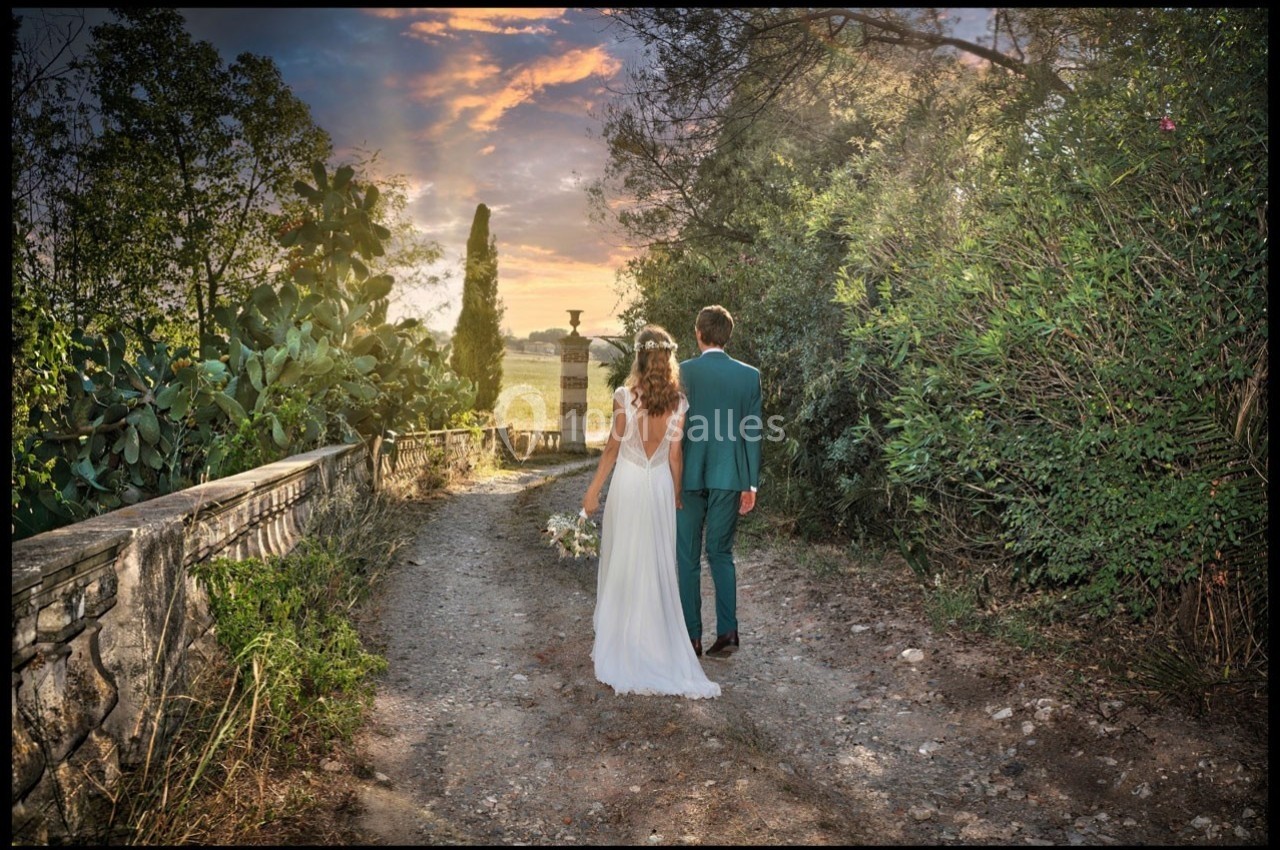 Un couple en tenue de mariage marche sur un chemin bordé de végétation, sous un ciel coloré au coucher du soleil.