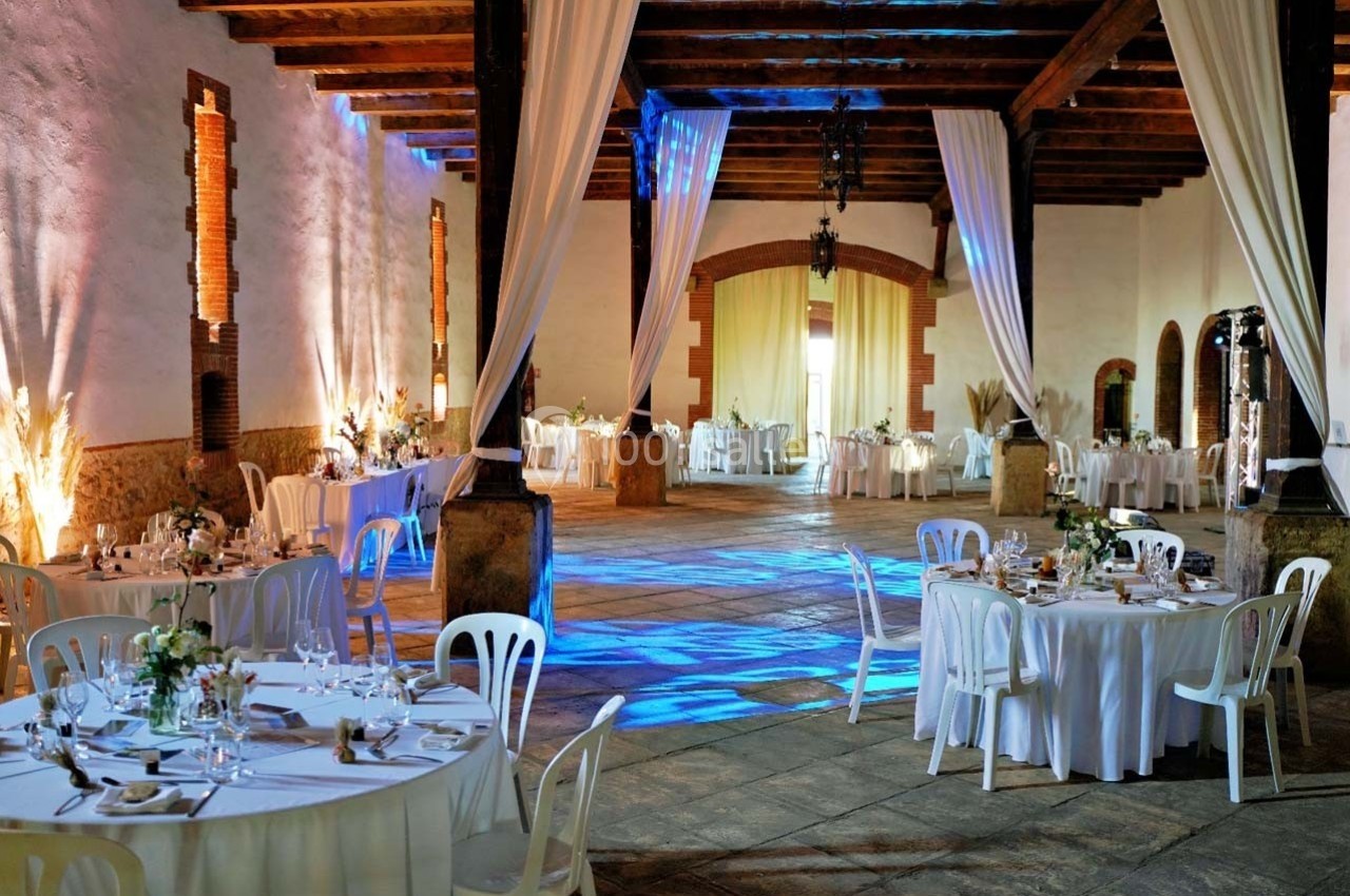 Salle de réception spacieuse avec tables rondes dressées, éclairage coloré et rideaux blancs suspendus.