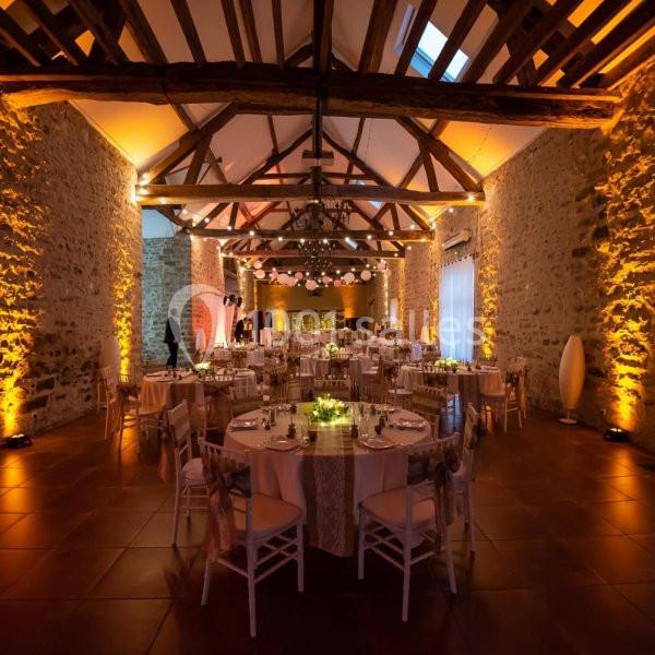 Salle de réception avec murs en pierre, poutres apparentes, tables décorées et éclairage chaleureux.