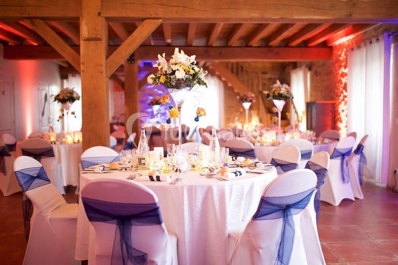 Salle de réception décorée avec des tables rondes, nappes blanches, chaises ornées de nœuds bleus et centres de table…