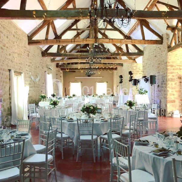 Salle de réception lumineuse avec des tables rondes décorées, des chaises blanches et une charpente en bois apparente.