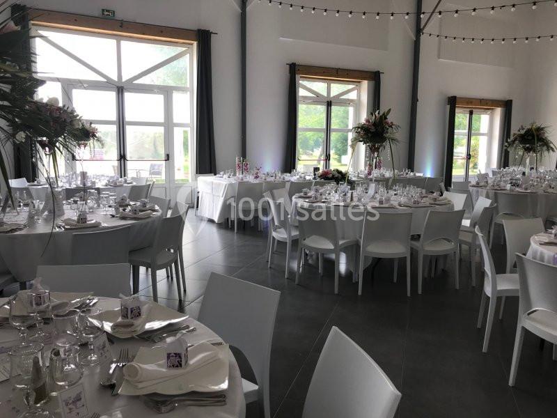 Salle de réception lumineuse avec tables rondes décorées pour un événement, chaises blanches et grandes fenêtres.