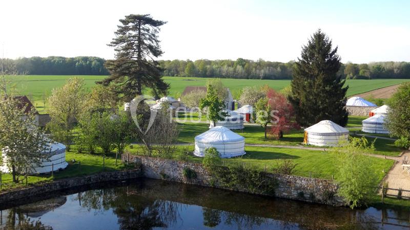 Vue d'un ensemble de yourtes blanches installées dans un paysage verdoyant avec des arbres et un étang au premier plan.