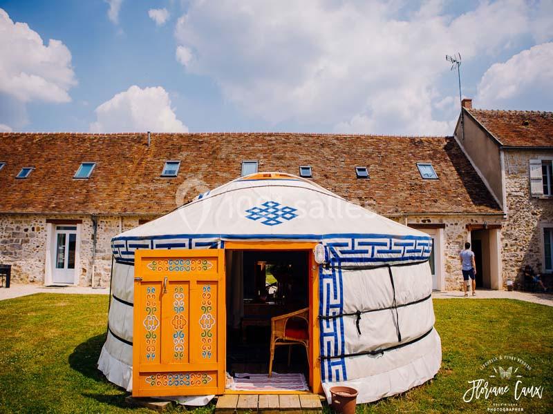 Yourte blanche décorée de motifs bleus et orange, installée devant une maison en pierre sous un ciel partiellement nuageux.