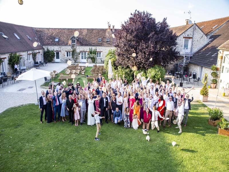 Groupe de personnes rassemblées dans une cour verdoyante devant des bâtiments en pierre, levant les bras en l'air.