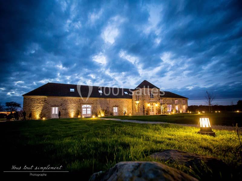 Bâtiment en pierre éclairé à la tombée de la nuit, entouré d'une pelouse et d'un ciel nuageux.