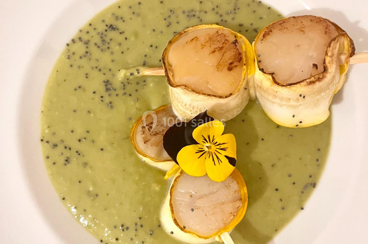 Brochettes de noix de Saint-Jacques sur un lit de velouté vert parsemé de graines, décorées d'une fleur comestible.