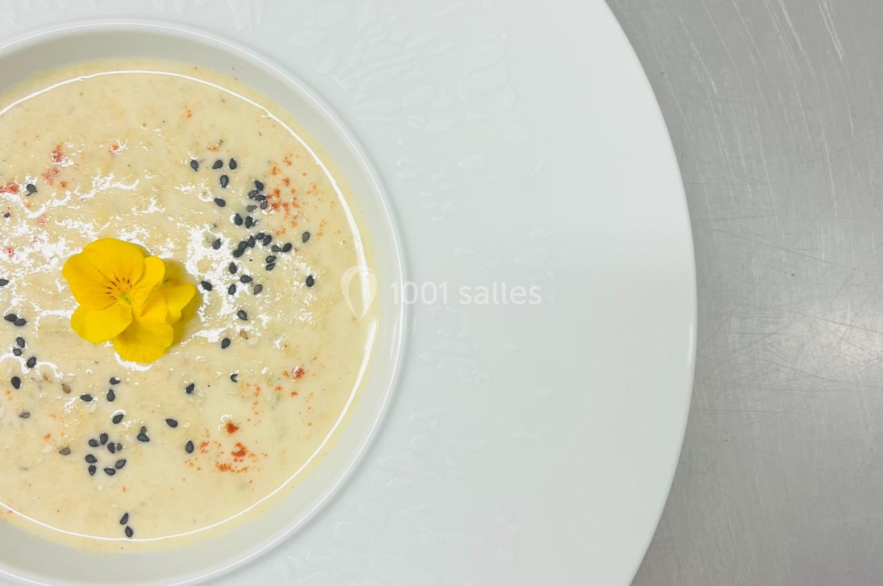 Soupe crémeuse servie dans une assiette blanche, décorée d'une fleur jaune et de graines noires.