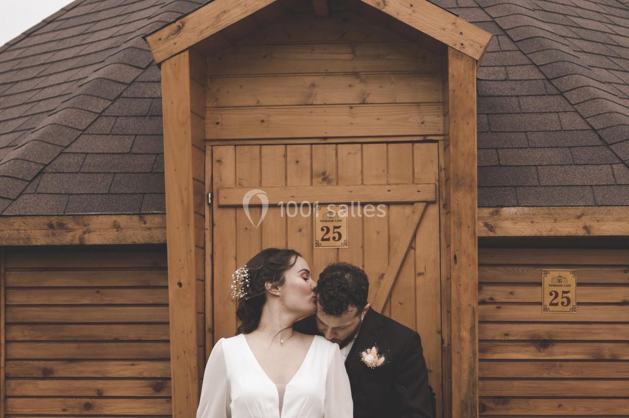 Un couple en tenue de mariage pose devant une petite cabane en bois avec un toit en pente.