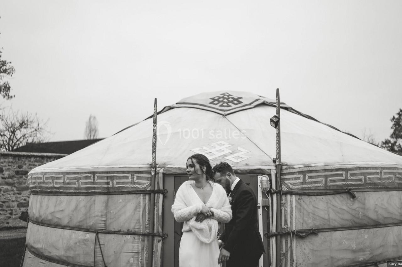 Un couple en tenue de mariage se tient devant une yourte dans un décor extérieur sobre et nuageux.