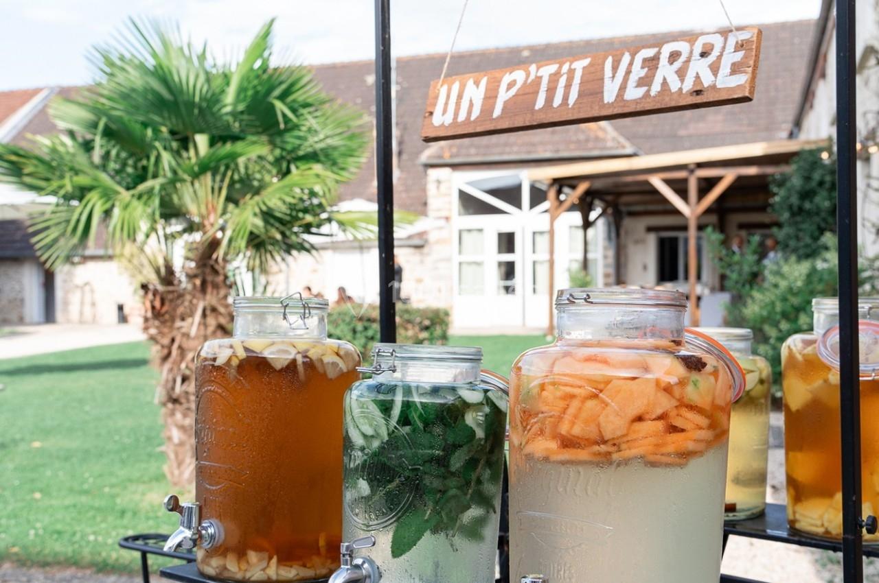 Des bonbonnes de boissons fruitées et infusées exposées en extérieur devant un bâtiment avec une enseigne ’Un p'tit verre’.