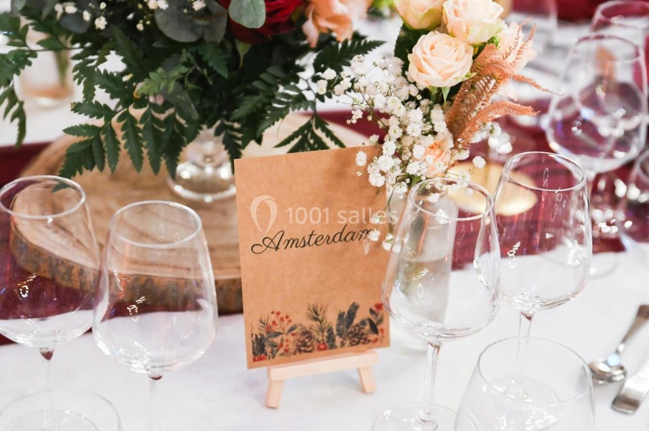 Carte ’Amsterdam’ sur un chevalet entourée de fleurs, verres à vin et décorations de table élégantes.