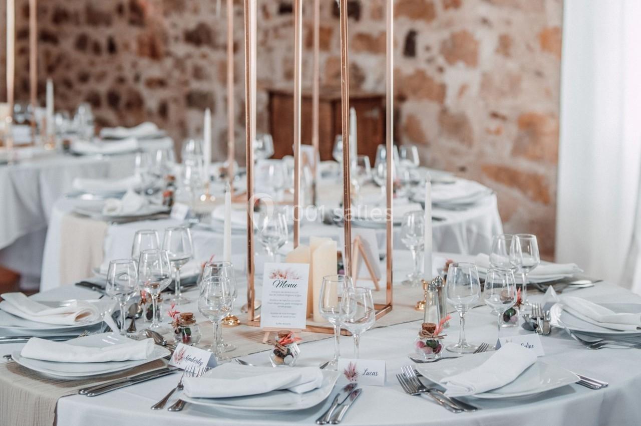 Tables élégamment dressées pour un événement, avec nappes blanches, bougies et décorations florales dans une salle en pierre.