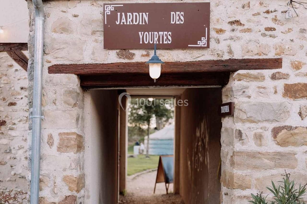 Entrée voûtée en pierre avec une enseigne ’Jardin des Yourtes’, menant à un espace extérieur avec une yourte visible.