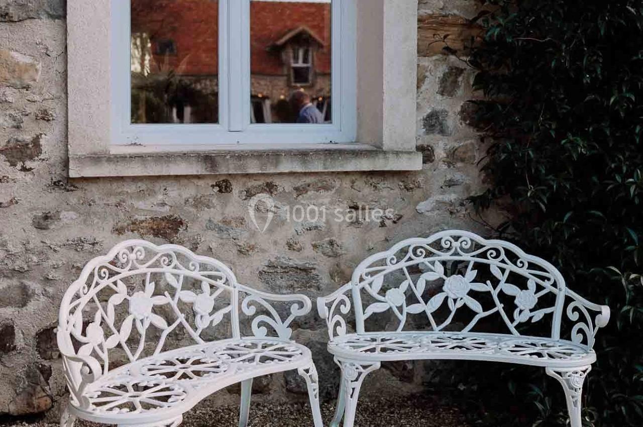 Deux bancs en fer forgé blanc ornés de motifs floraux, placés devant un mur en pierre avec une fenêtre.