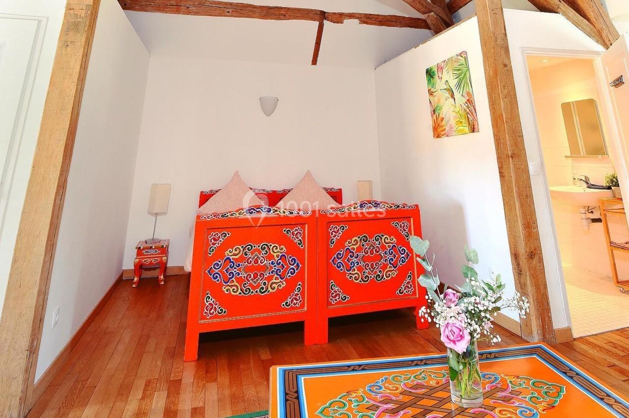 Lit en bois peint de motifs colorés dans une chambre lumineuse avec poutres apparentes et bouquet de fleurs sur une table.