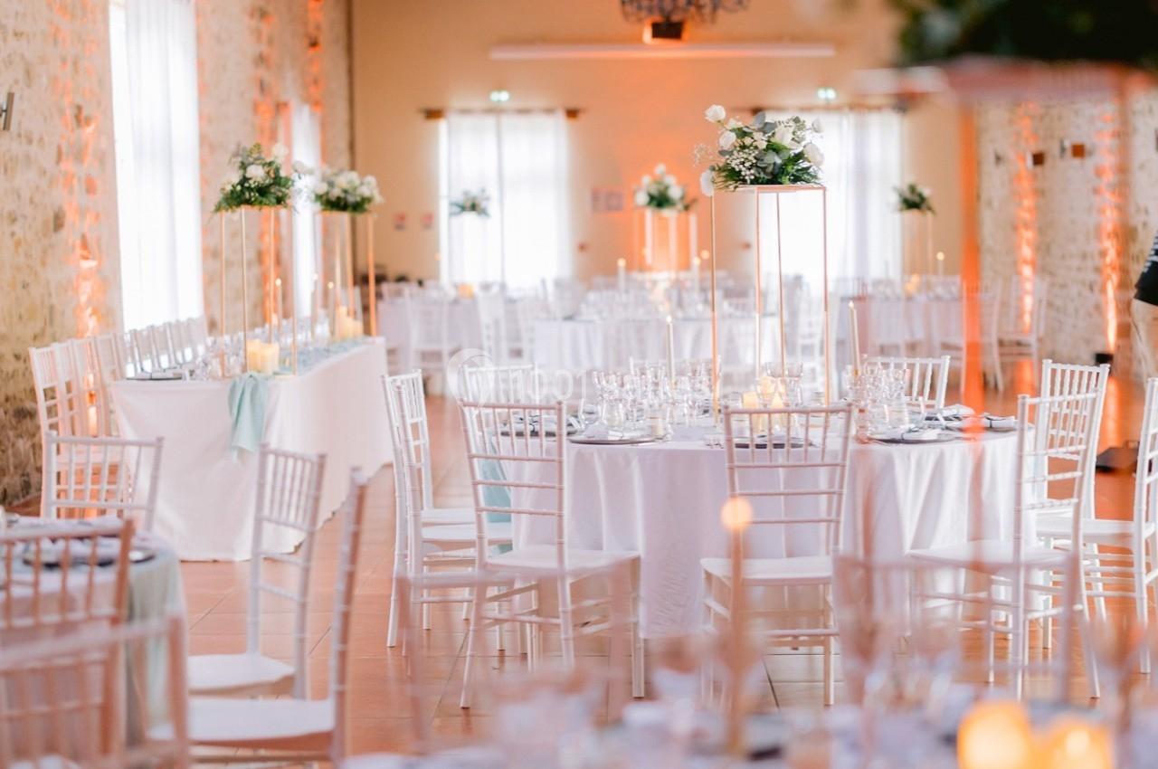 Salle de réception décorée avec des tables rondes, nappes blanches, chaises blanches et centres de table fleuris.