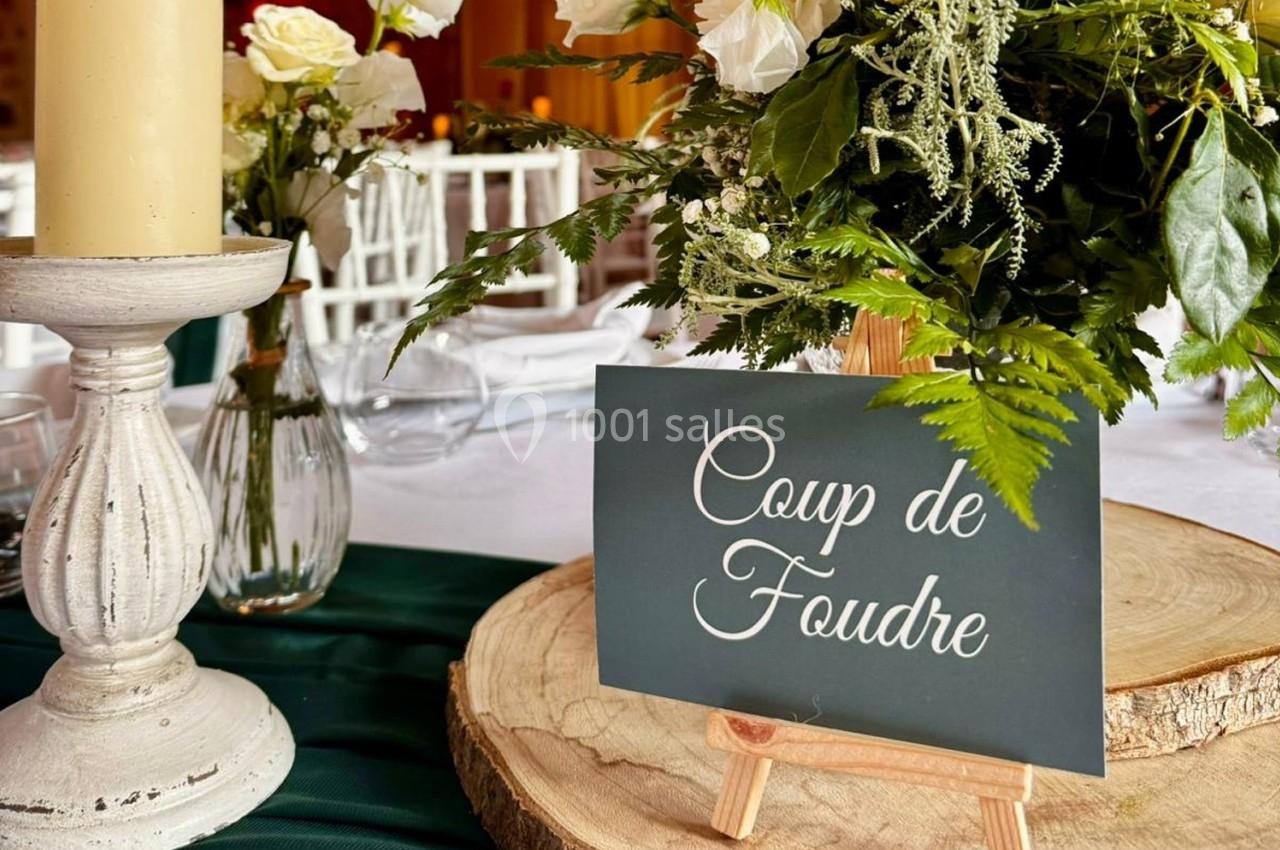 Carte ’Coup de Foudre’ posée sur un chevalet en bois, entourée de fleurs et d'une bougie sur une table décorée.