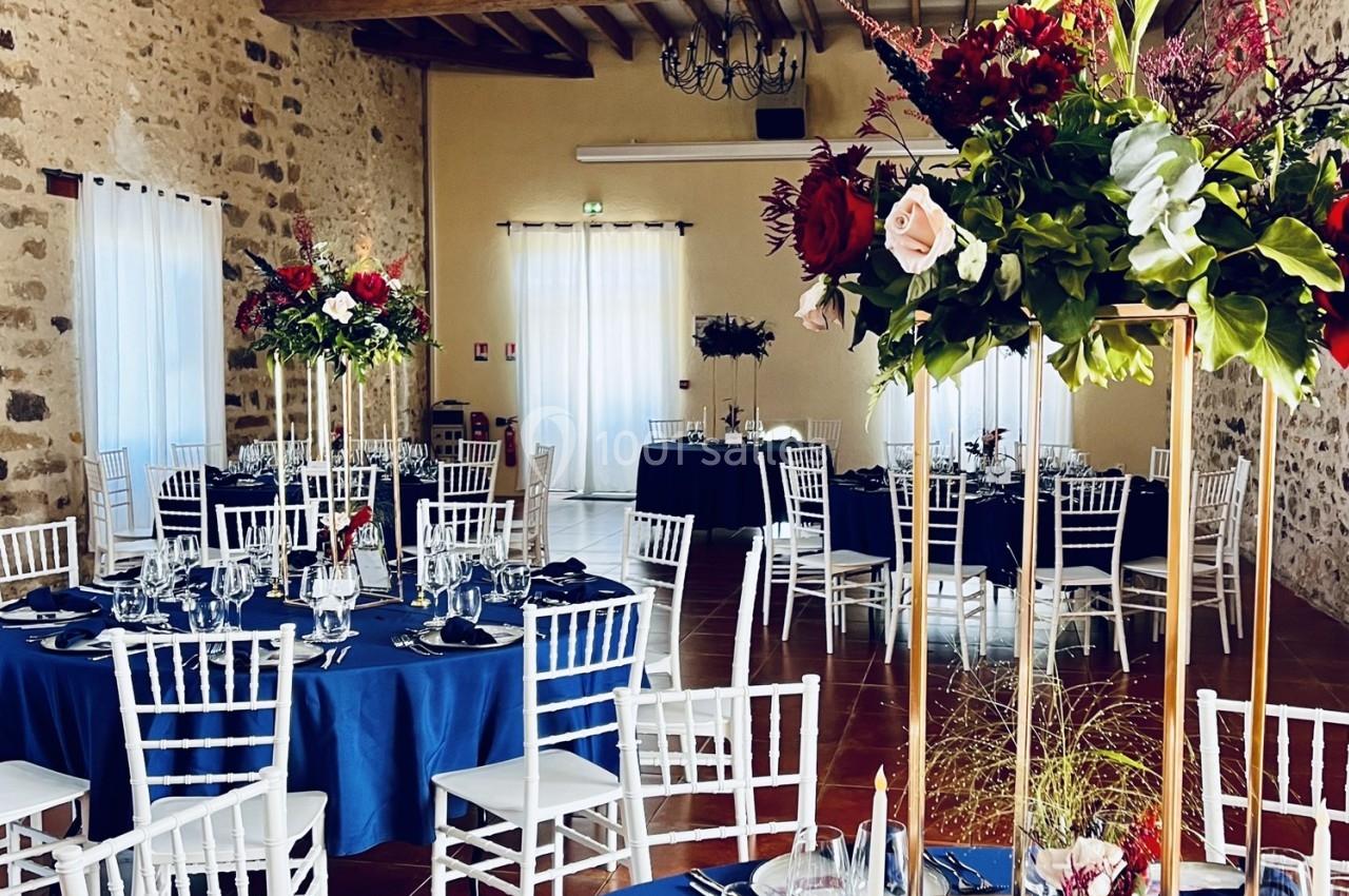Salle de réception décorée avec des tables rondes, nappes bleues, chaises blanches et centres de table fleuris.