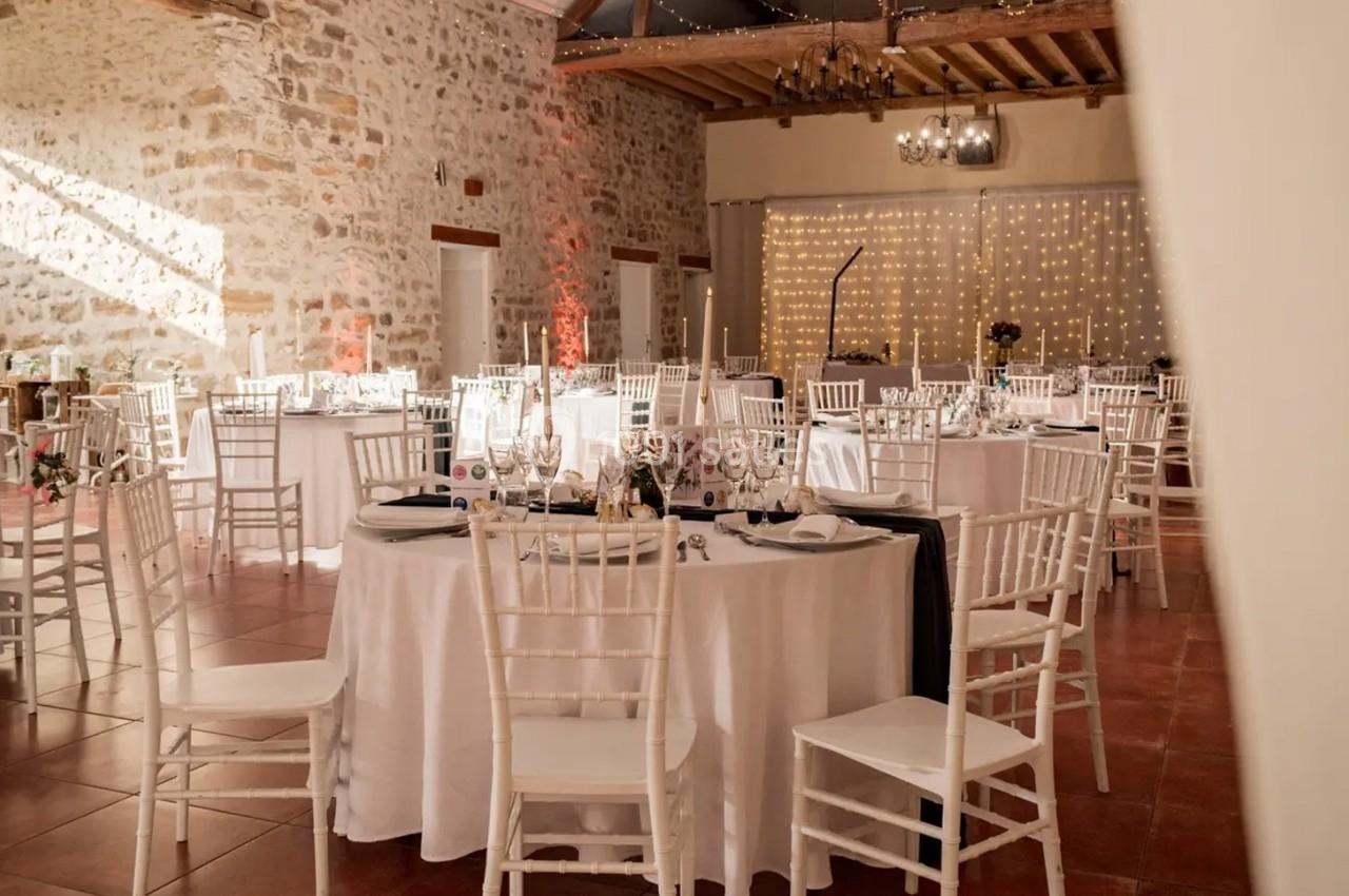 Salle de réception décorée avec des tables rondes dressées, chaises blanches et guirlandes lumineuses sur un mur en pierre.