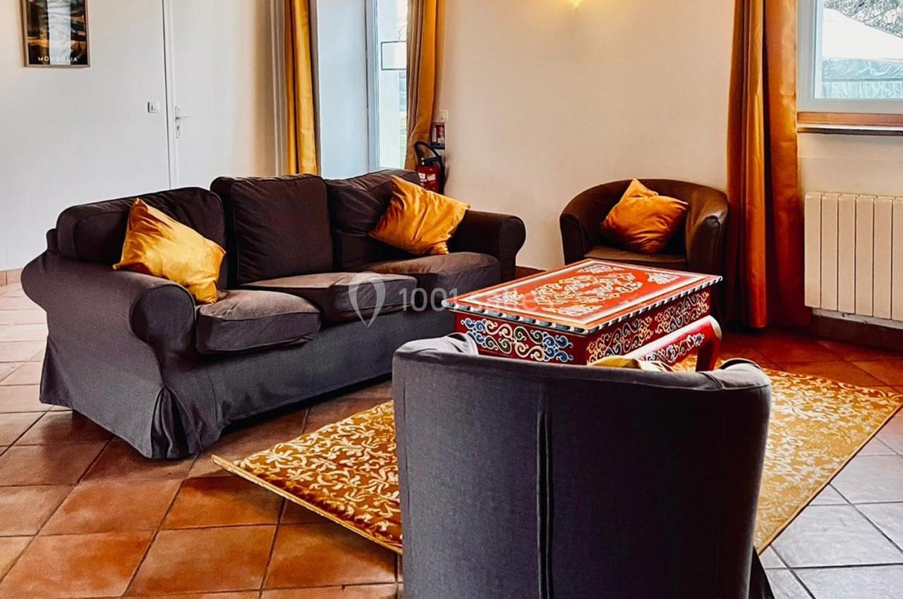 Salon avec canapé et fauteuils gris, coussins orange, table basse décorée et tapis assorti sur un sol en carrelage.