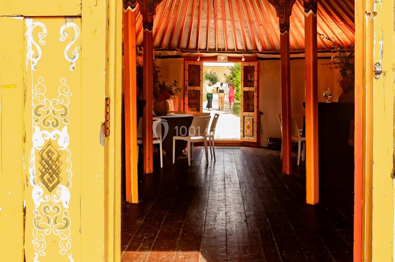 Vue intérieure d'une tente colorée avec des tables et des chaises, ouverte sur un jardin ensoleillé à l'arrière-plan.