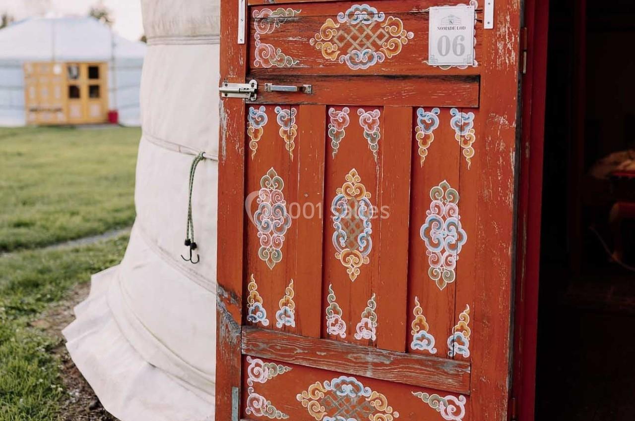 Porte en bois rouge ornée de motifs traditionnels, ouverte sur une yourte blanche dans un cadre herbeux.