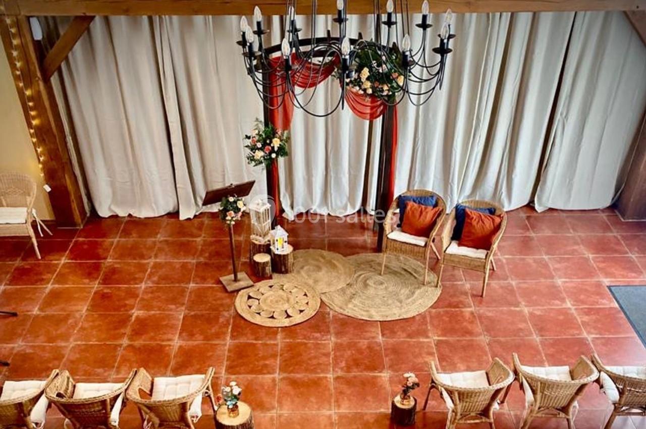 Salle décorée pour une cérémonie avec chaises en rotin, tapis, fleurs et un lustre suspendu au plafond.