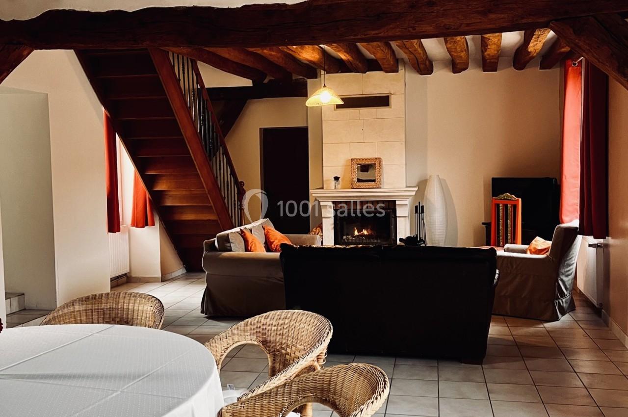 Salon chaleureux avec poutres apparentes, cheminée allumée, canapés, table en osier et escalier en bois.