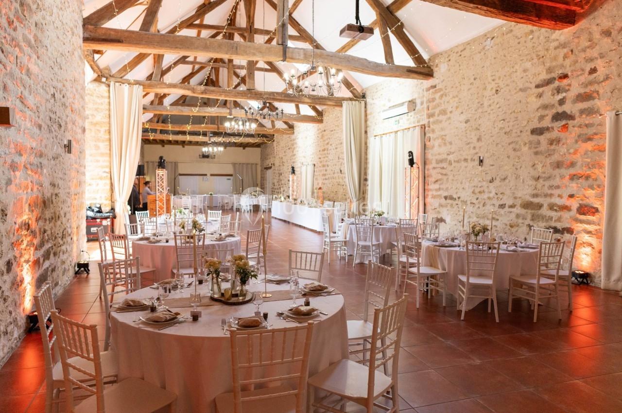 Salle de réception lumineuse avec murs en pierre, poutres apparentes et tables rondes décorées pour un événement.