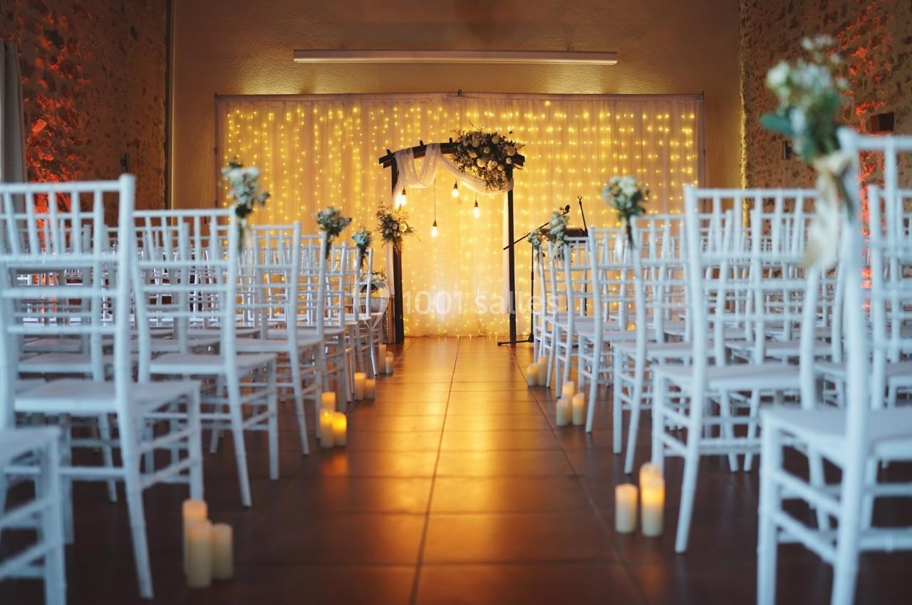 Allée centrale décorée de chaises blanches, bougies et guirlandes lumineuses menant à une arche florale.