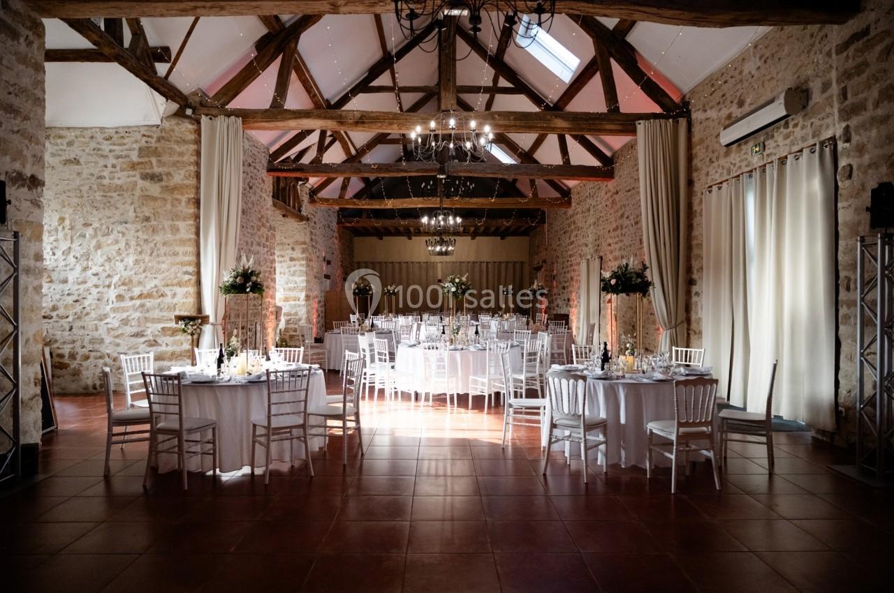 Salle de réception en pierres apparentes avec tables rondes dressées et décorations florales sous un plafond en poutres…