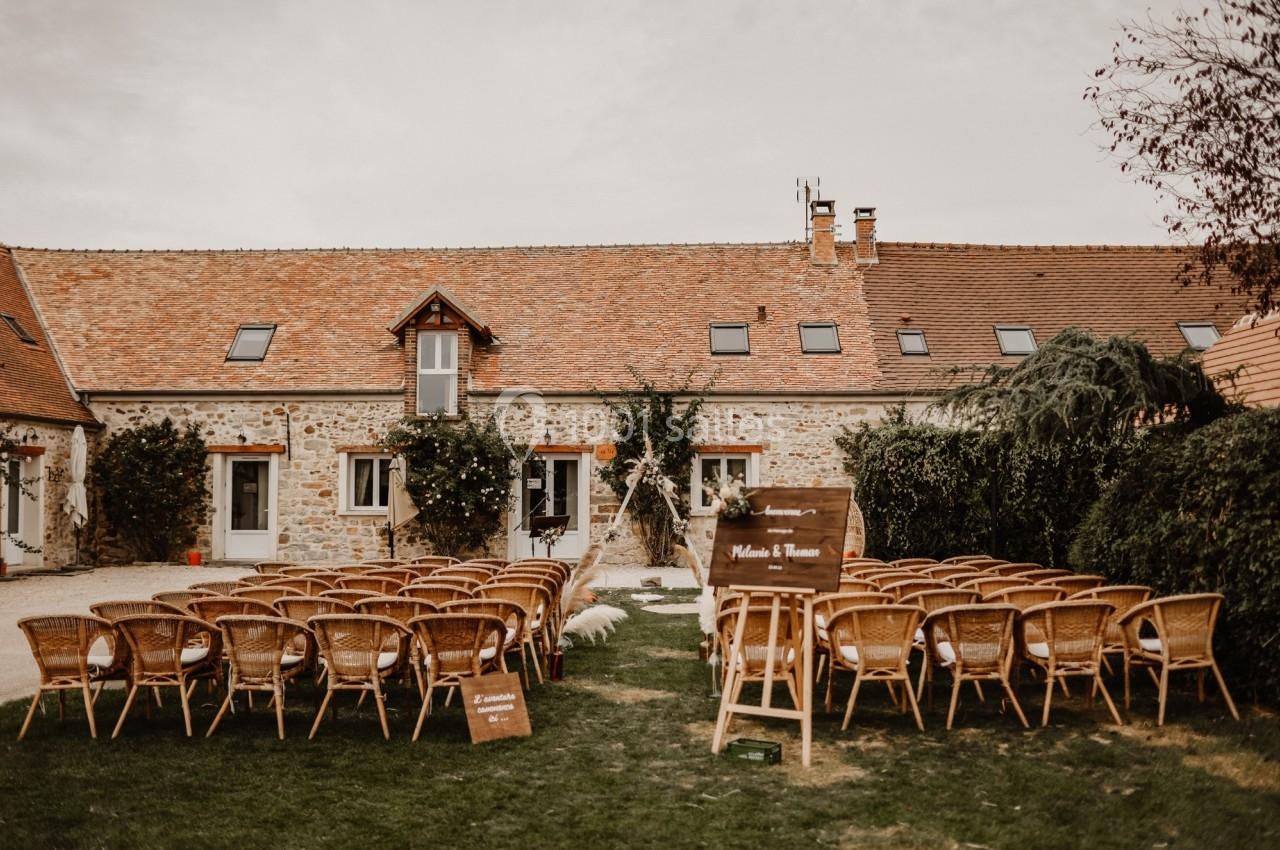Chaises en rotin disposées en extérieur devant une maison en pierre pour une cérémonie avec arche décorée.