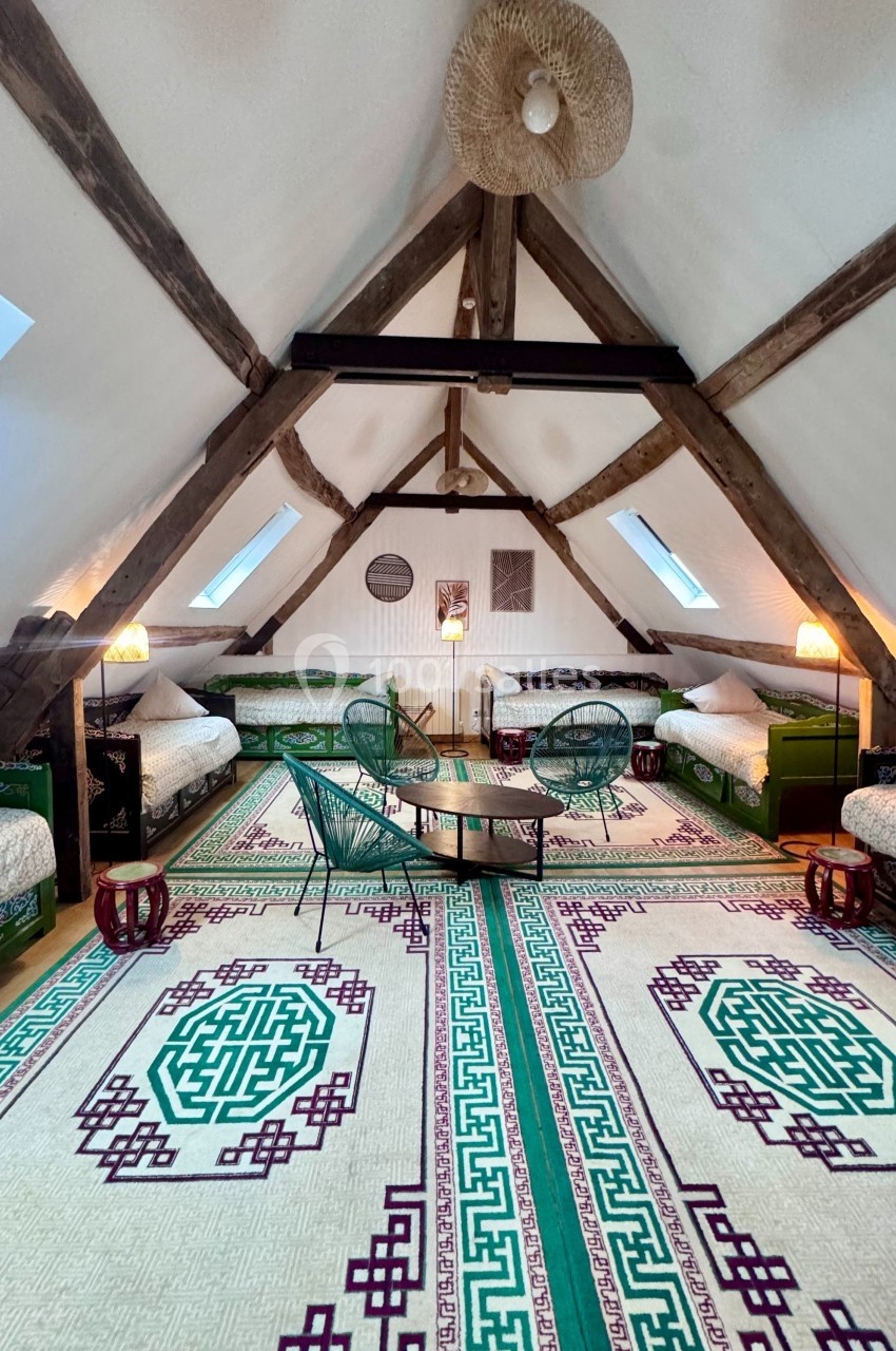 Grenier aménagé avec poutres apparentes, tapis coloré, lits simples, fauteuils en osier et éclairage chaleureux.