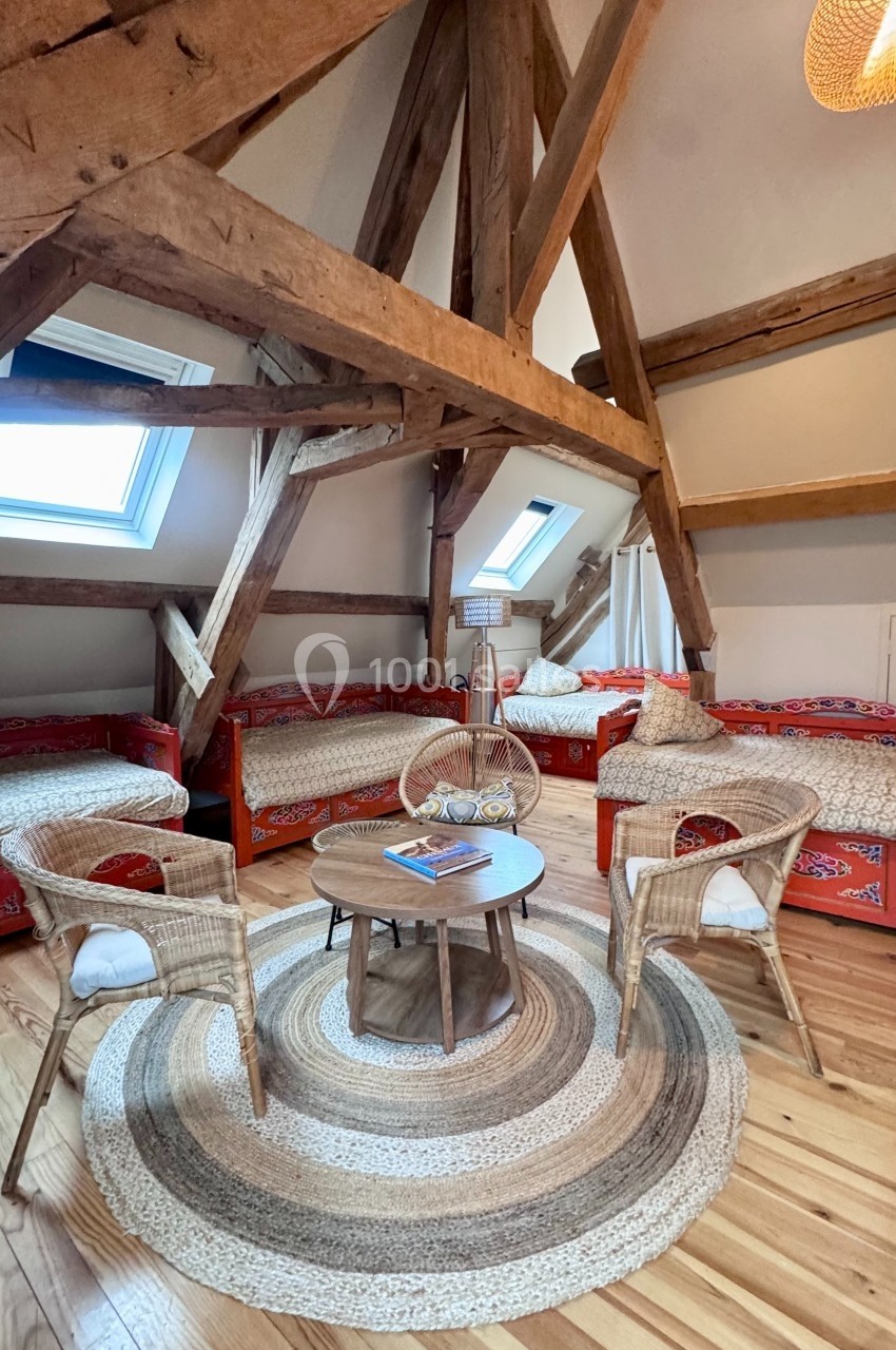 Salon mansardé avec poutres apparentes, fauteuils en rotin, tapis rond et lits banquettes aux motifs colorés.