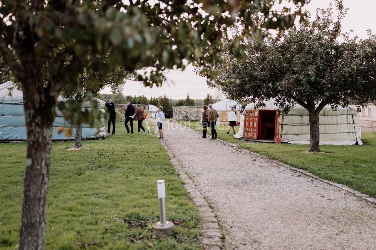 Chemin bordé d'arbres menant à des yourtes dans un espace vert, avec des personnes se promenant.