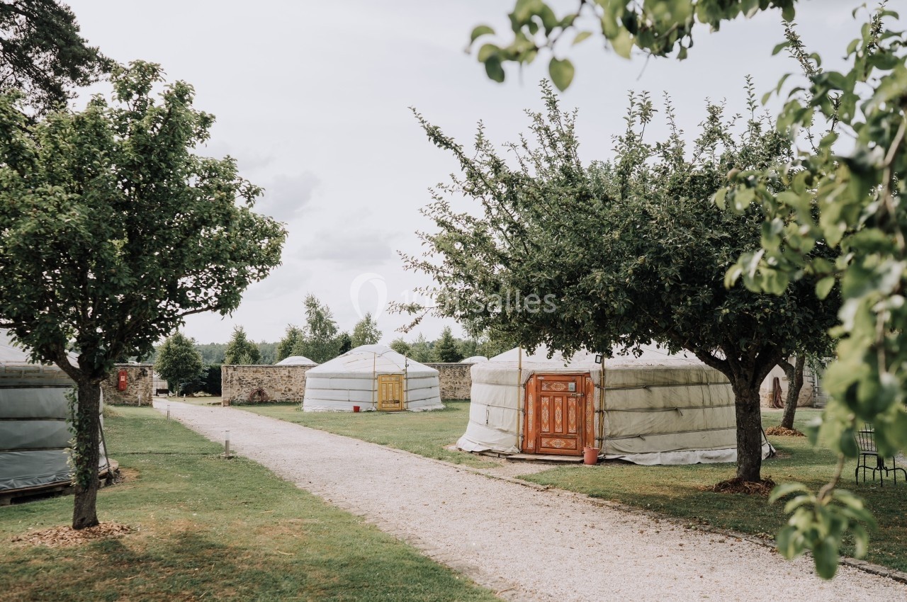 Plusieurs yourtes blanches avec portes en bois, disposées dans un jardin verdoyant avec des arbres et un chemin gravillonné.