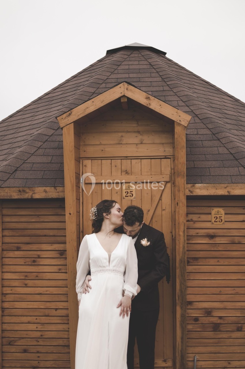 Un couple en tenue de mariage pose devant une petite cabane en bois avec un toit en pente.