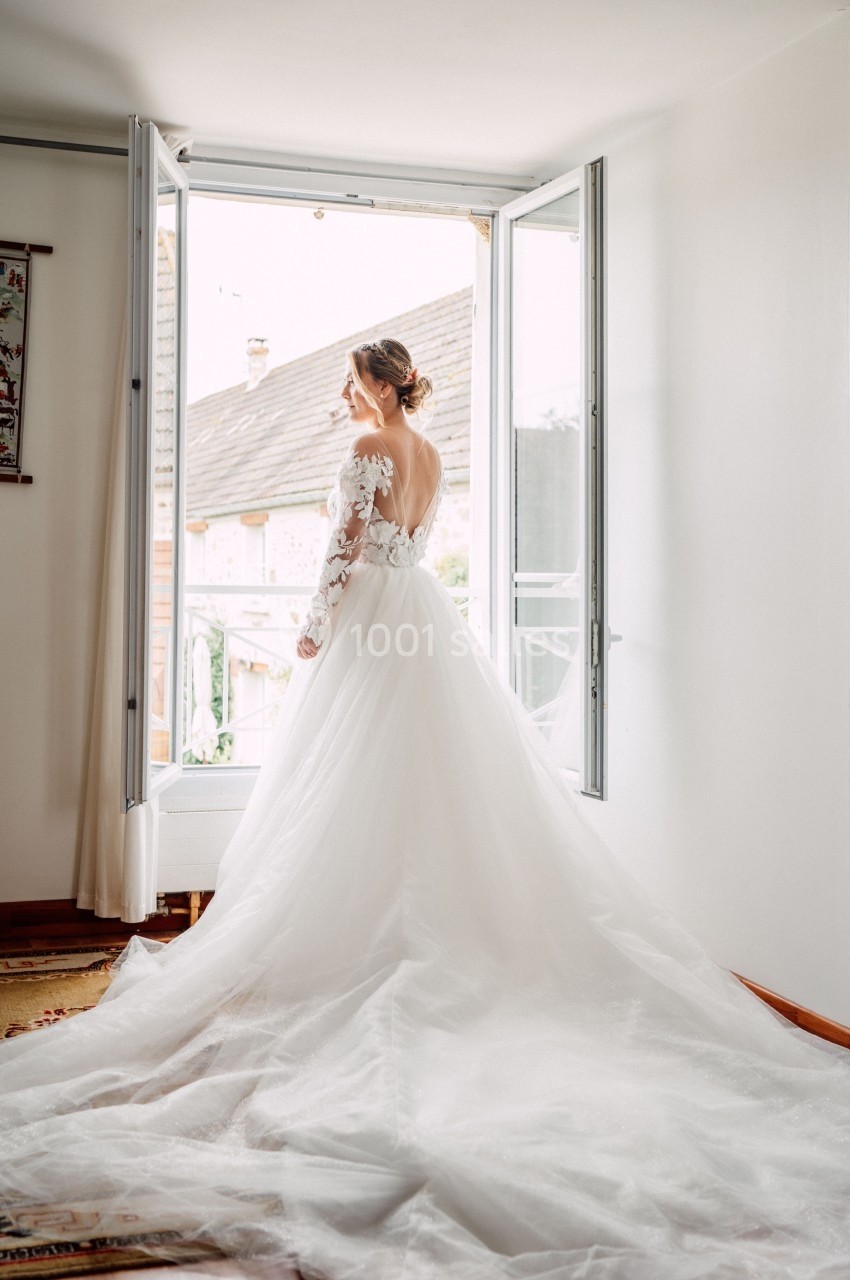 Femme en robe de mariée blanche, vue de dos, regardant par une fenêtre ouverte dans une pièce lumineuse.
