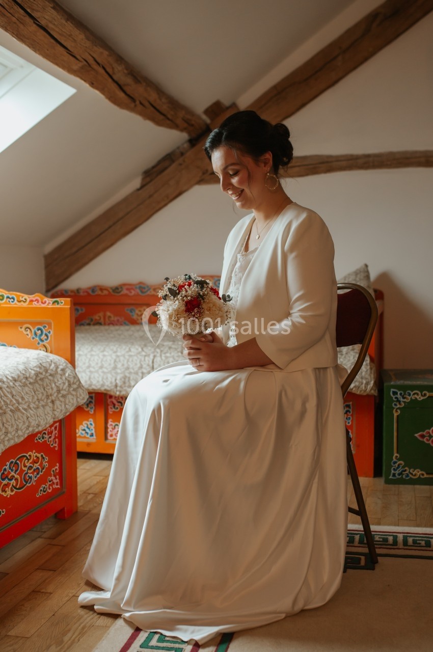 Femme en robe blanche assise sur une chaise, tenant un bouquet de fleurs, dans une pièce au décor traditionnel coloré.