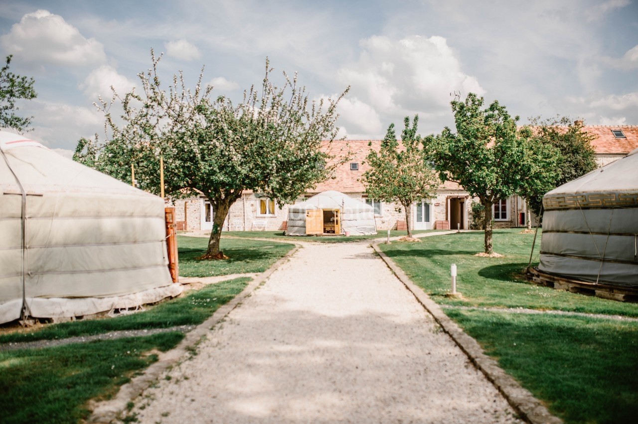 Allée centrale bordée de yourtes et d'arbres, menant à un bâtiment en pierre sous un ciel partiellement nuageux.
