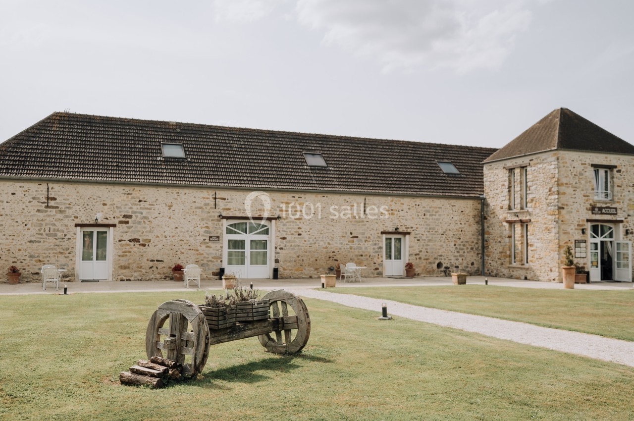 Bâtiment en pierre avec toit en tuiles, fenêtres blanches et une charrette en bois sur une pelouse.